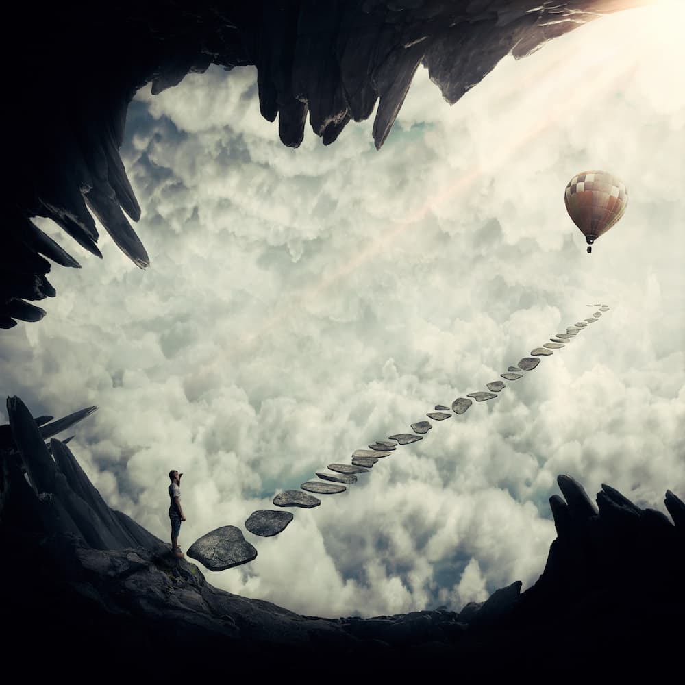 A person on a cliff edge looking at stepping stones leading through the clouds toward a hot-air balloon